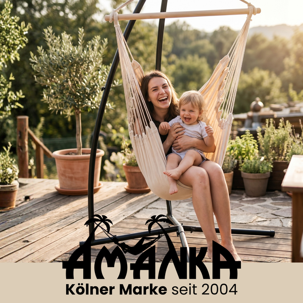 Eine Frau und ein Kind sitzen lachend in einem beigen Stoff-Hängesessel mit schwarzem Metallgestell auf einer sonnigen Terrasse. AMANKA Logo.