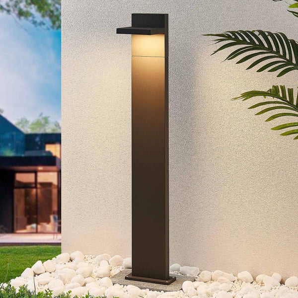 Moderne rechteckige Außen-Sockelleuchte in Anthrazit, warmes Licht nach unten strahlend, auf weißen Kieselsteinen vor einer strukturierten Wand.