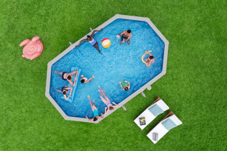 Draufsicht auf einen ovalen Aufstellpool im Garten mit schwimmenden Personen, Luftmatratze, Wasserball und Sonnenliegen auf dem Rasen.