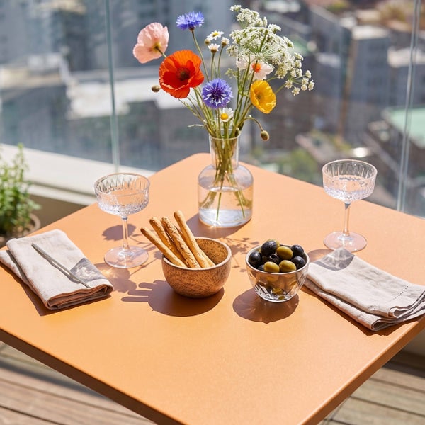Gedeckter quadratischer Balkontisch mit Wildblumen in einer Glasvase, Schalen mit Oliven und Grissini sowie zwei Gläsern auf einer Dachterrasse.