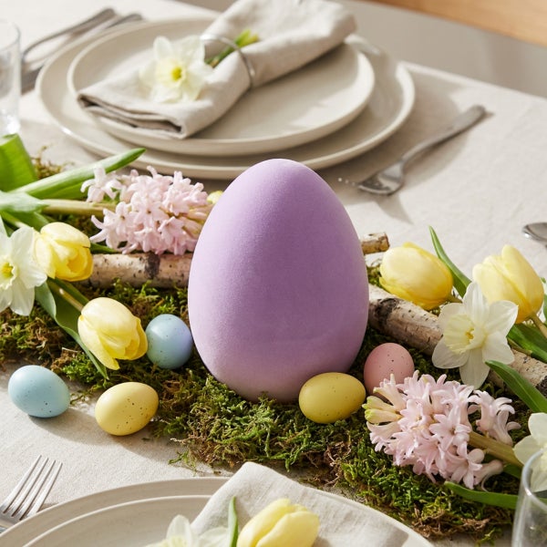 Osterdekoration mit einem großen violetten Osterei, umgeben von Moos, Tulpen, Narzissen, Hyazinthen und kleinen Ostereiern auf einem gedeckten Tisch.