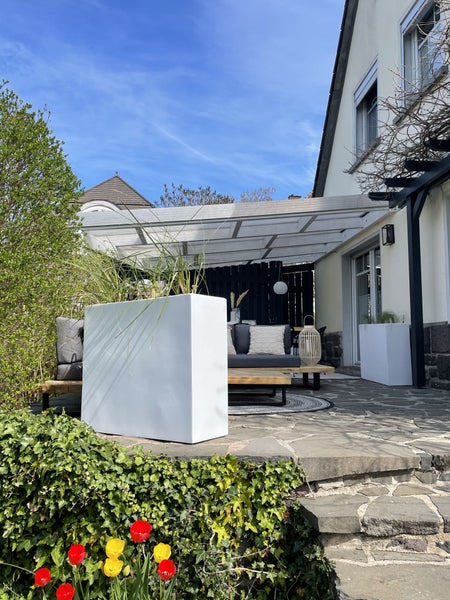 Modern gestaltete Terrasse mit weißem Pflanzkasten, Holzgartentisch, schwarzen Flechtstühlen, Pergola und hängenden Papierlampions.