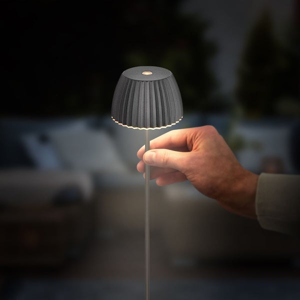 Eine Hand hält den Stab einer kleinen dunkelgrauen LED-Tischleuchte mit plissiertem Schirm in einer dunklen Umgebung.
