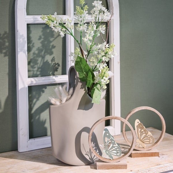 Dekorative Vase aus Keramik mit kunstlichen Blumen und zwei hölzernen Schmetterlingsaufstellern als Frühlingsdekoration auf einem Holztisch.