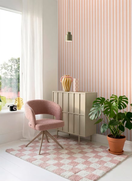 Hell gestalteter Raum mit gestreifter Vliestapete, rosa Polstersessel, beige Kommode, kariertem Teppich und einer Monstera-Zimmerpflanze.