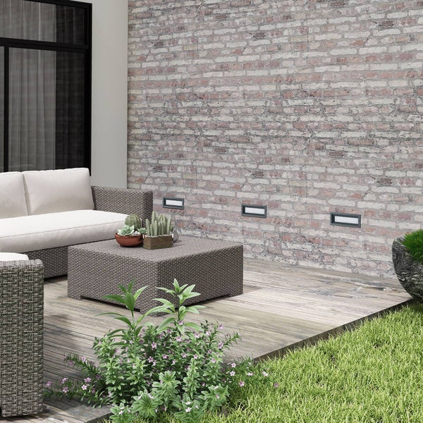 Drei rechteckige LED-Wandeinbauleuchten in einer Wand in Backsteinoptik auf einer modernen Terrasse mit Polyrattan-Möbeln und Holzdielen.