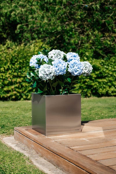 Quadratischer Pflanzkübel aus Edelstahl mit blauen Hortensien auf einer Holzterrasse im Garten.