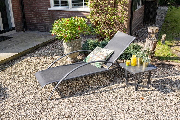 Moderne graue Sonnenliege mit Metallgestell und Textilgewebe neben passendem Beistelltisch auf Kies in einem hellen Garten.