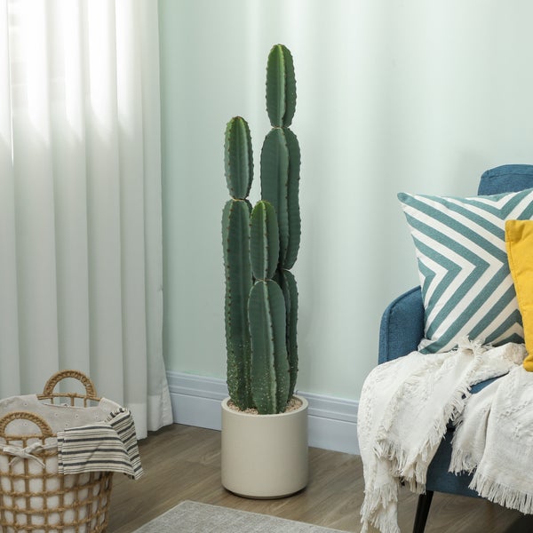 Künstlicher Kaktus in einem beigen Topf in einem hellen Wohnzimmer neben einem blauen Sessel mit dekorativen Kissen und einem Flechtkorb.