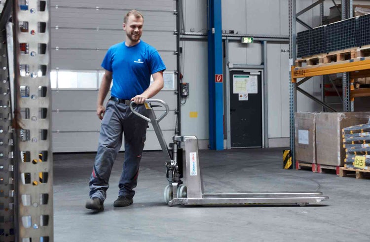 Ein Mitarbeiter bedient einen Hubwagen aus Edelstahl in einem hellen, organisierten Lager mit Hochregalen und Paletten.