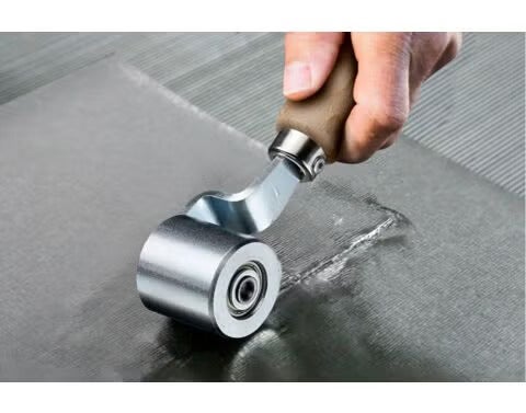 Anpressrolle aus Metall mit Holzgriff bei der Bearbeitung einer Naht auf einer grauen Fläche.