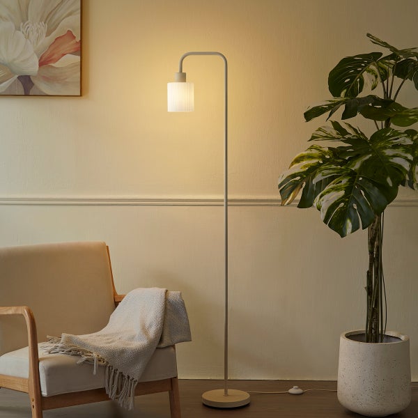 Minimalistische Stehleuchte mit Metallgestell und geriffeltem Glasschirm in einem hellen Wohnzimmer mit Sessel und Monstera-Pflanze.