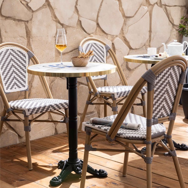Bistro-Set mit runden Tischen in Marmoroptik und geflochtenen Stühlen auf einer Holzterrasse vor einer Natursteinwand.