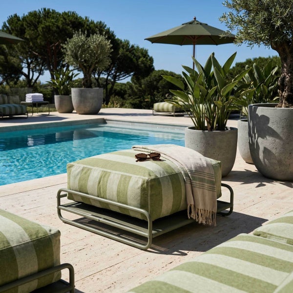 Garten-Pouf mit grün gestreiftem Kissen auf Metallgestell an einem Pool, umgeben von Pflanzen und Sonnenschirmen auf einer hellen Terrasse.