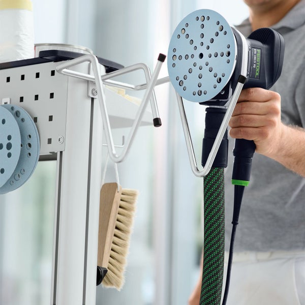 Festool Exzenterschleifer in einem Werkzeughalter aus Metall an einem Werkstattwagen. Daneben hängen Schleifblätter und eine Holzhandbürste.