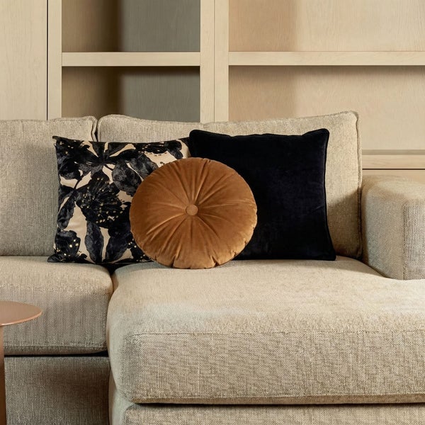 Drei Dekokissen auf einem hellen Sofa, ein quadratisches Kissen mit Blumenmuster, ein rundes braunes Samtkissen und ein schwarzes Kissen.