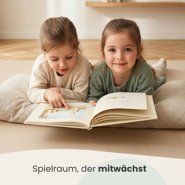 Zwei Kinder liegen auf einem großen Bodenkissen auf einer strukturierten Spielmatte und lesen gemeinsam ein Buch in einem hellen Zimmer.