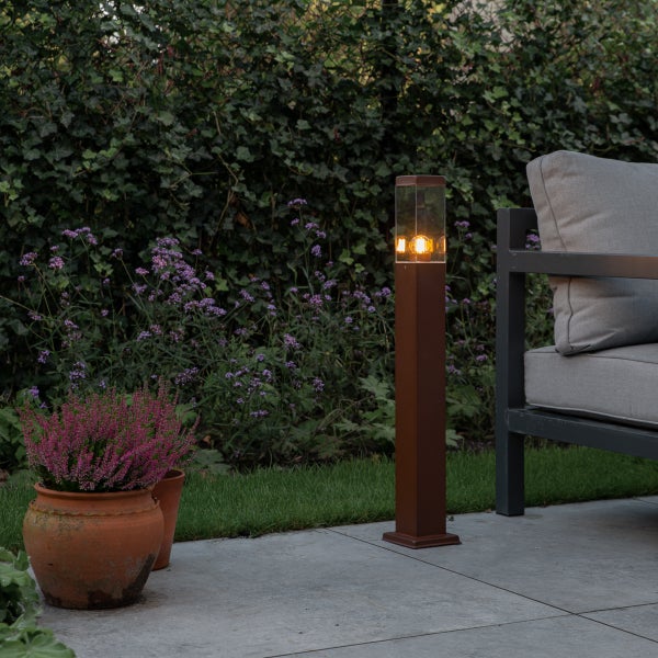 Rechteckige Wegeleuchte in Rostoptik mit Glaseinsatz auf einer Steinterrasse neben einem Gartenstuhl und einer Pflanze im Topf vor einer Hecke.