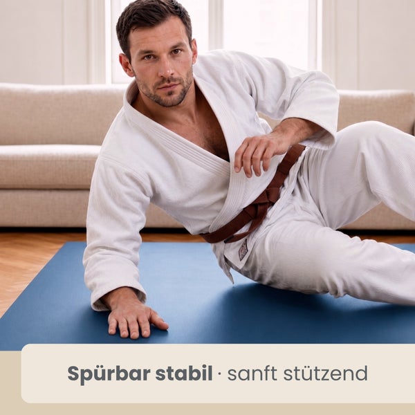 Ein Mann in einem Kampfsportanzug macht Bodenübungen auf einer stabilen und stützenden Fitnessmatte in einem hellen Wohnzimmer.