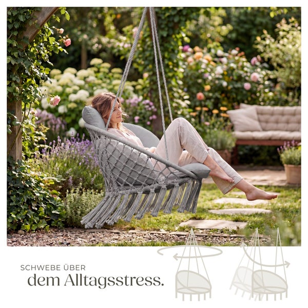 Eine Frau entspannt in einem grauen Makramee-Hängesessel mit Kissen in einem blühenden Garten mit Blumen und einer Holzbank.