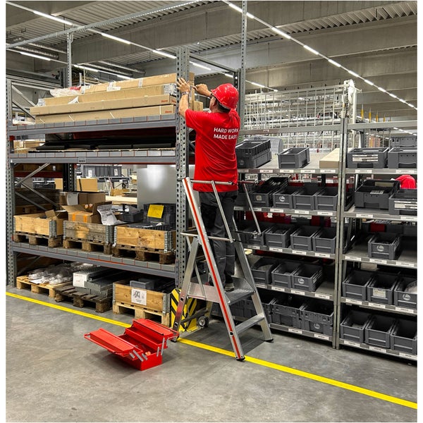 Ein Mann arbeitet mit einer Leiter in einem Lagerregal, Werkzeugkasten steht daneben.