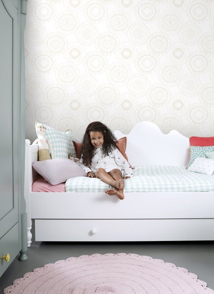Kind sitzt auf einem weißen Bett in einem hellen Raum mit gemusterter Tapete.