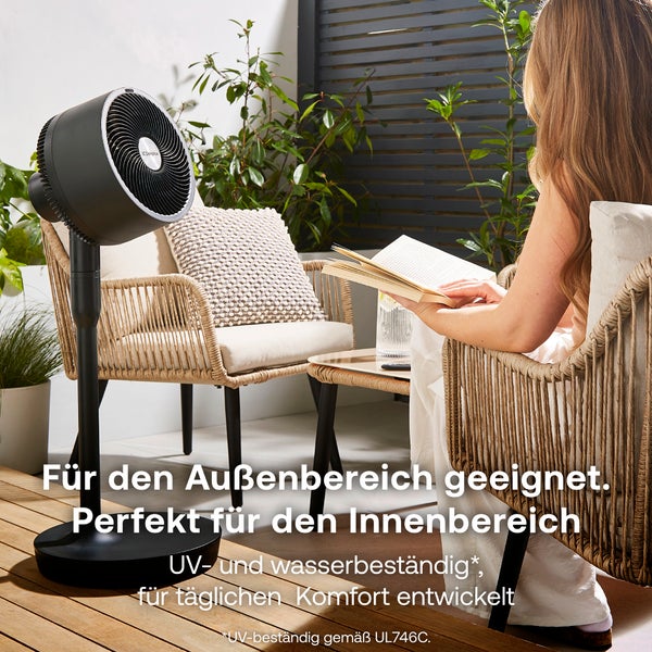 Schwarzer Standventilator auf einer Holzterrasse neben einer Person im Korbstuhl, wasserbeständig und für den Innen- und Außenbereich geeignet.