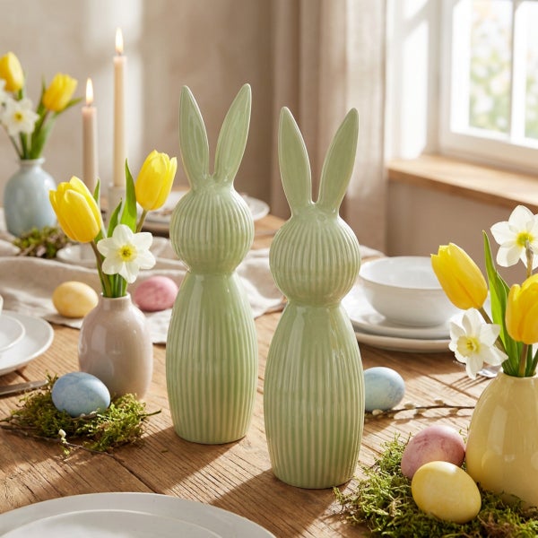Zwei dekorative Osterhasen aus Keramik mit Rillenstruktur als Tischdekoration auf einem Esstisch mit Blumenvasen und Ostereiern.