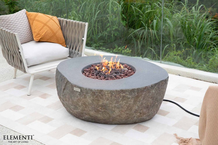 Elementi Gaskamin Boulder in Naturstein-Optik auf einer Terrasse mit Sessel und Pflanzen im Hintergrund.