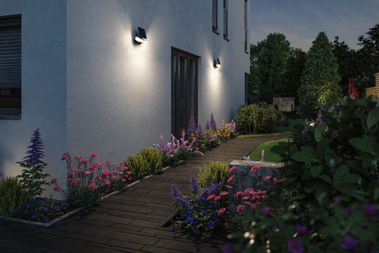 Moderner Garten mit zwei Solar-LED-Wandstrahlern, die einen gepflasterten Weg und Blumenbeete an einer Hausfassade beleuchten.