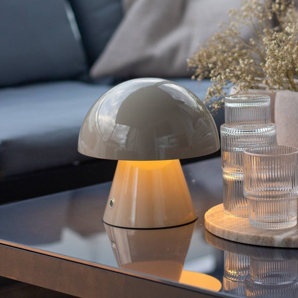 Akkubetriebene Tischleuchte in Pilzform auf einem gläsernen Gartentisch neben Gläsern und Blumen in gemütlicher Abendstimmung.