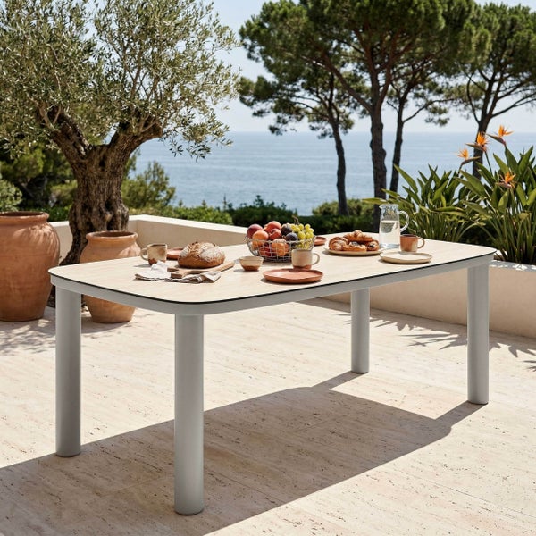 Moderner Gartentisch auf einer sonnigen Terrasse mit Meerblick, gedeckt mit Frühstück, umgeben von Olivenbäumen und Terrakotta-Töpfen.