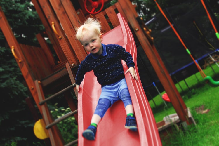 Kleinkind auf einer roten Rutsche an einem Spielturm aus Holz im Garten.