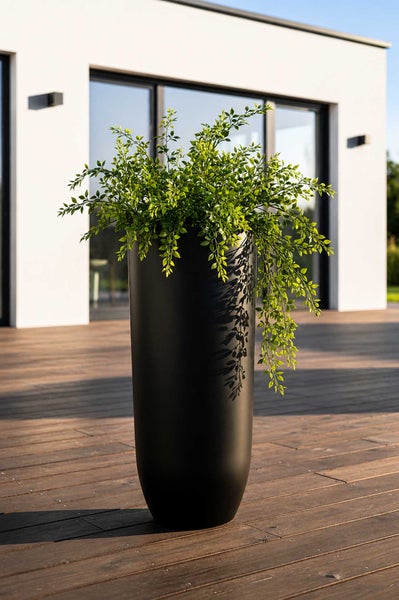 Hoher, zylindrischer Pflanzkübel in Schwarz mit einer grünen Pflanze auf einer Holzterrasse vor einem modernen Haus.