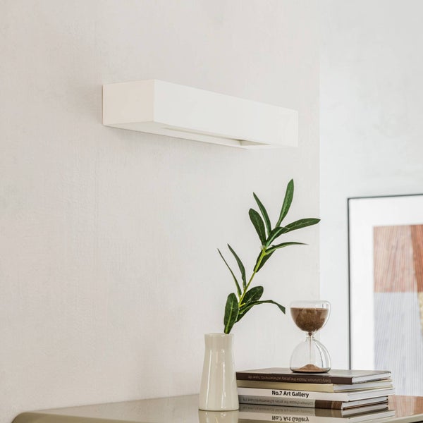 Moderne rechteckige Wandleuchte in Weiß an einer hellen Wand über einer Vase mit Pflanze und Büchern in minimalistischem Design.