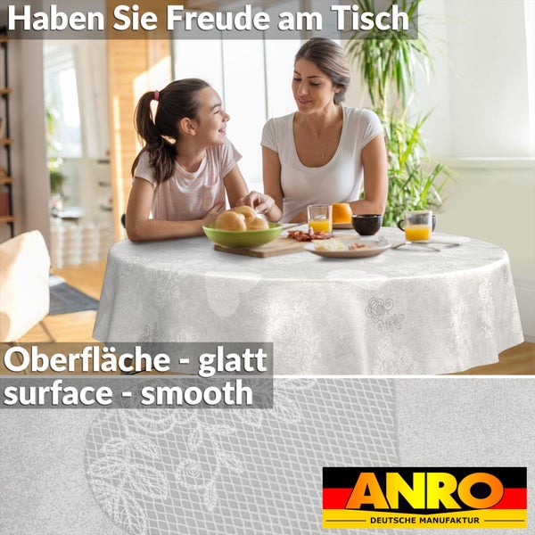 Runde, weiße, abwaschbare Tischdecke mit floralem Spitzenmuster und glatter Oberfläche in heller Wohnszene. ANRO DEUTSCHE MANUFAKTUR Logo.