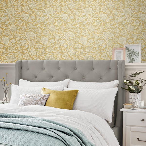 Schlafzimmer mit gepolstertem Kopfteil und floraler Mustertapete an der Wand, kombiniert mit Textilien und Nachttisch.