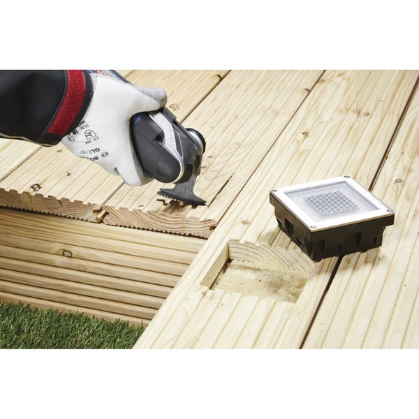 Eine Person verwendet ein Multifunktionswerkzeug, um ein Loch in eine Holzterrasse zu schneiden, neben der eine quadratische Bodeneinbauleuchte liegt.