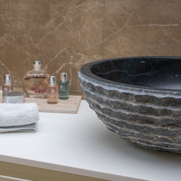 Modernes Badezimmer mit runden Aufsatzwaschbecken aus dunklem Naturstein mit strukturierter Außenseite auf weißer Platte.