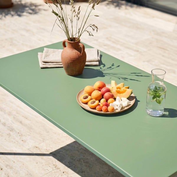 Grüner Gartentisch auf einer hellen Terrasse, dekoriert mit einem Obstteller, einer Terrakottavase mit Gräsern und einer Wasserkaraffe.