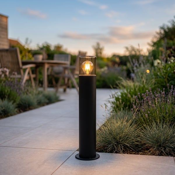Schwarze Wegeleuchte aus Metall mit Glaszylinder und sichtbarem Leuchtmittel auf einem hellen Gartenweg zwischen Pflanzen.