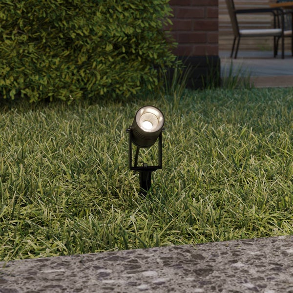 Eingeschalteter LED-Gartenstrahler mit Erdspieß auf einer Rasenfläche vor einem Busch.