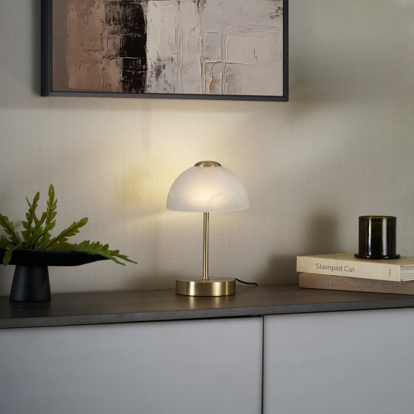 Moderne Tischleuchte mit pilzförmigem weißem Glasschirm und Messingfuß auf einem Sideboard neben einer Pflanze und Büchern.