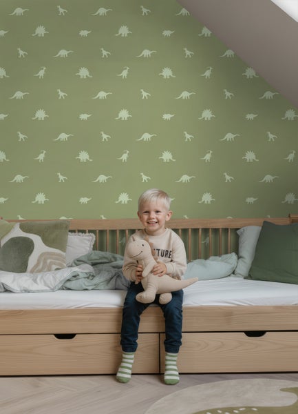 Hell eingerichtetes Kinderzimmer mit Vliestapete im Dinosaurier Muster. Ein Junge sitzt mit einem Stoff Dinosaurier auf einem Holzbett.