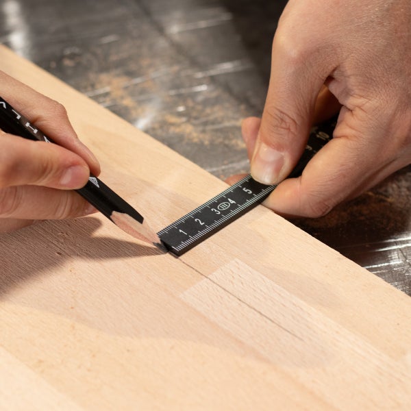 Eine Person markiert eine Holzplatte mit einem Bleistift und einem schwarzen Maßstab.