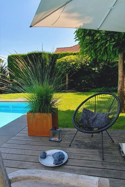 Moderner Garten mit Holzterrasse, Pool, schwarzem Loungesessel, Pflanzkübel in Edelrost-Optik mit Ziergras und kleiner Laterne.