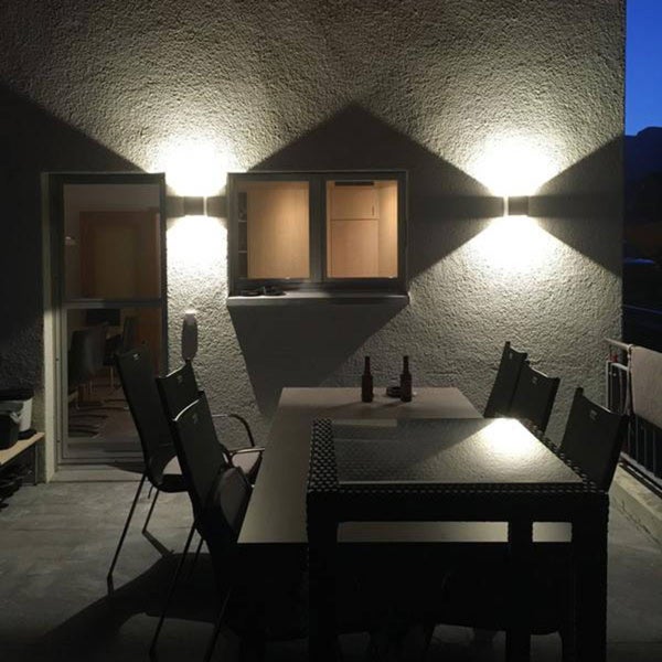 Moderne Außenwandleuchten an einer Fassade über einer Terrasse mit Tisch und Stühlen, die Licht nach oben und unten werfen.