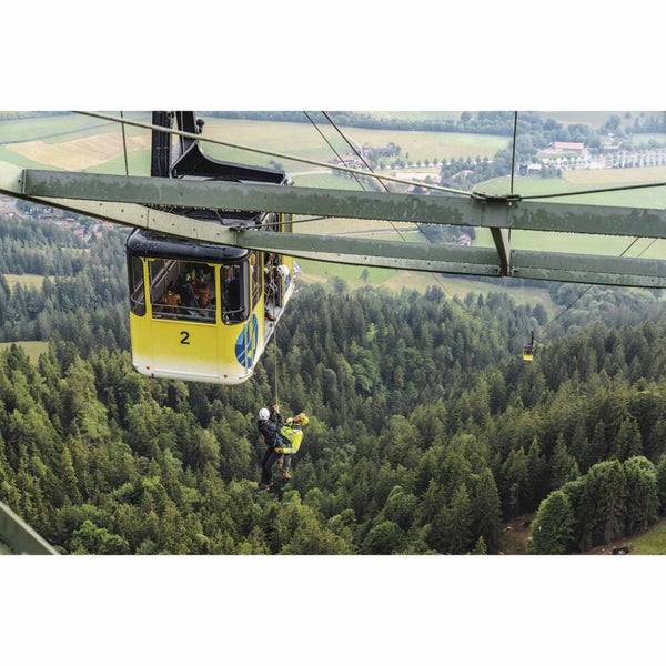 Zwei Personen in Sicherheitsausrüstung werden von einer gelben Seilbahnkabine über einem dichten Nadelwald abgeseilt.