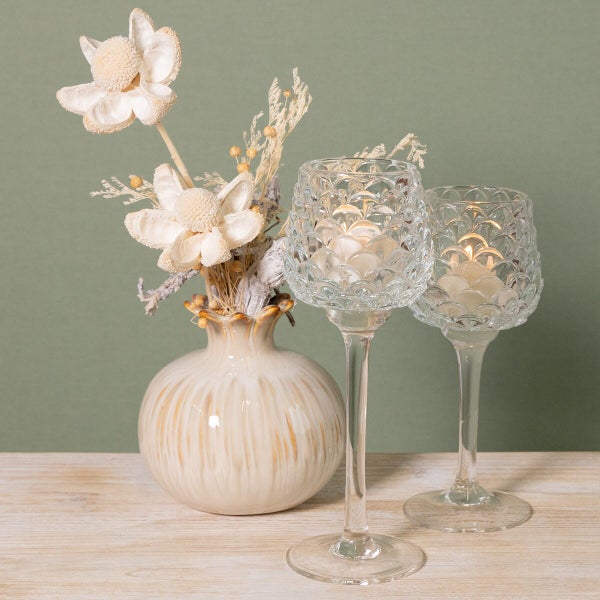 Zwei hohe Kerzenhalter aus strukturiertem Glas mit brennenden Teelichtern und eine Vase mit Trockenblumen auf einem Holztisch.
