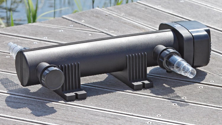 Schwarzer UVC-Teichklärer mit transparenten Schlauchanschlüssen auf einem Holzsteg am Teich.